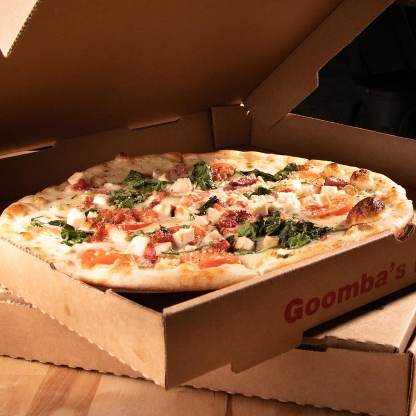 A Goomba's pizza with chicken, spinach, tomatoes, and bacon in an open delivery box.