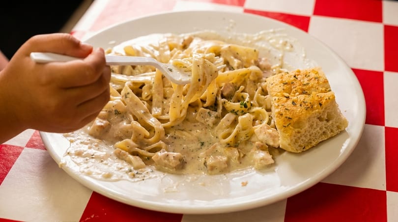 A hand twirls creamy chicken fettuccine alfredo with a fork, served with focaccia bread on a red and white checkered tablecloth.