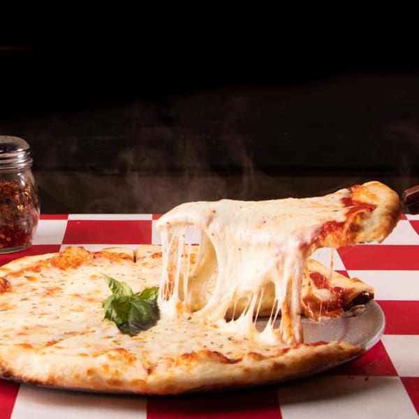 A hand lifts a slice of hot, cheesy pizza from a whole pie on a red and white checkered tablecloth.