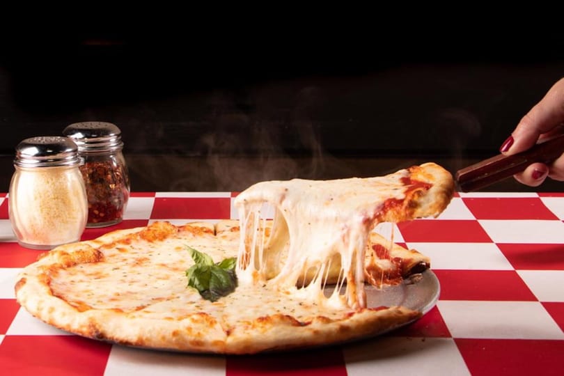 A hand lifts a slice of hot, cheesy pizza from a whole pie on a red and white checkered tablecloth.