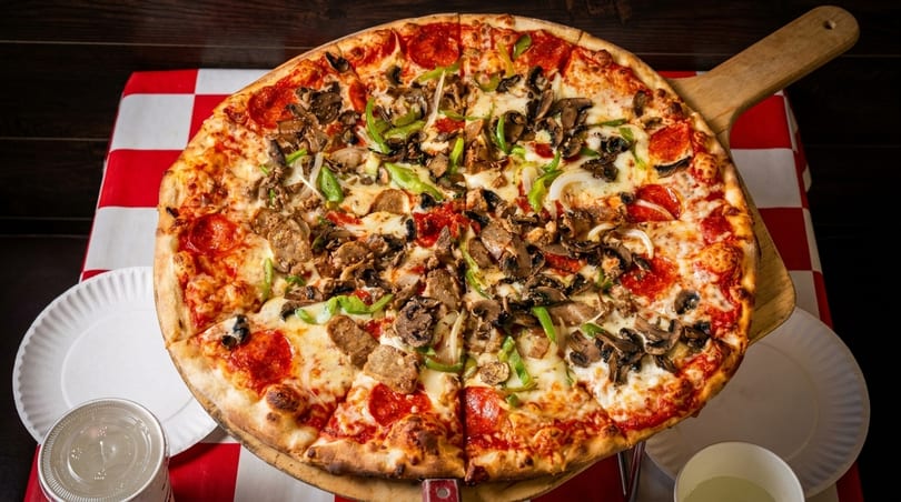 A large, sliced pizza with pepperoni, sausage, mushrooms, green peppers, and onions on a red and white checkered tablecloth.