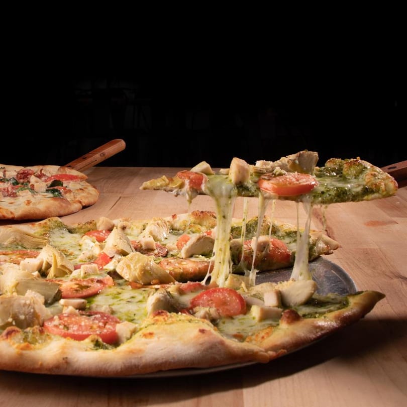 A slice of pesto chicken and artichoke pizza with melted cheese being lifted from a whole pizza.