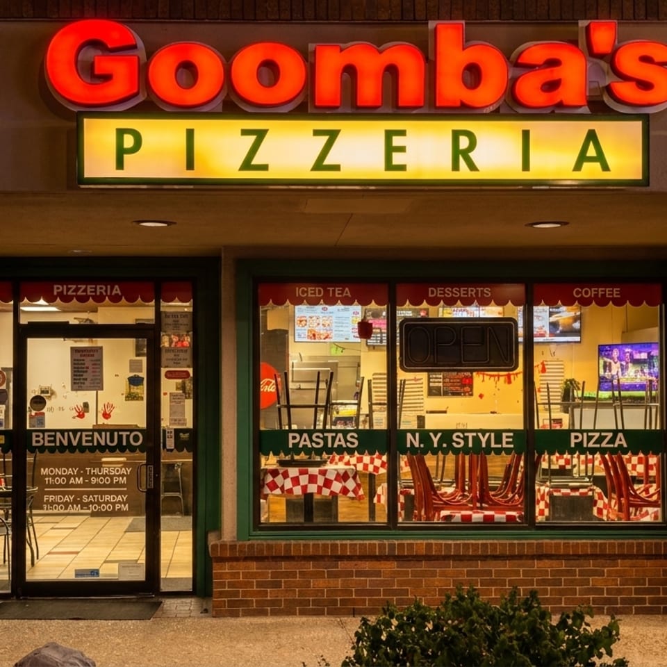 Exterior of Goomba's Pizzeria at night, with illuminated signs and visible interior dining area.