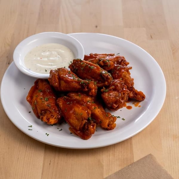 A white plate of buffalo chicken wings with ranch dressing on a light wooden table.