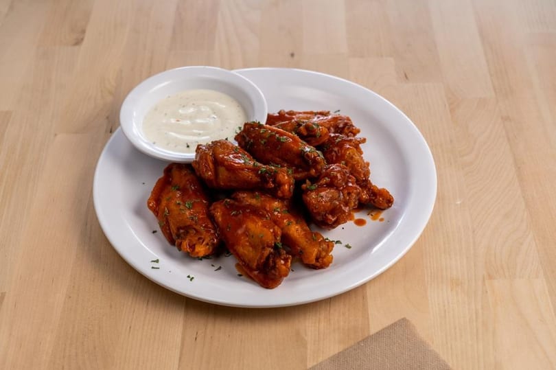 A white plate of buffalo chicken wings with ranch dressing on a light wooden table.