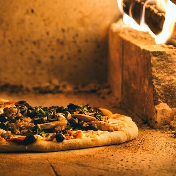 A fresh pizza with various toppings baking inside a wood-fired oven with glowing embers.